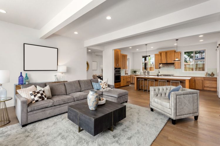 A stylish open concept living room seamlessly transitions into a kitchen area, featuring a large gray sectional sofa, white walls, wooden kitchen cabinetry, and a spacious layout with contemporary decor.